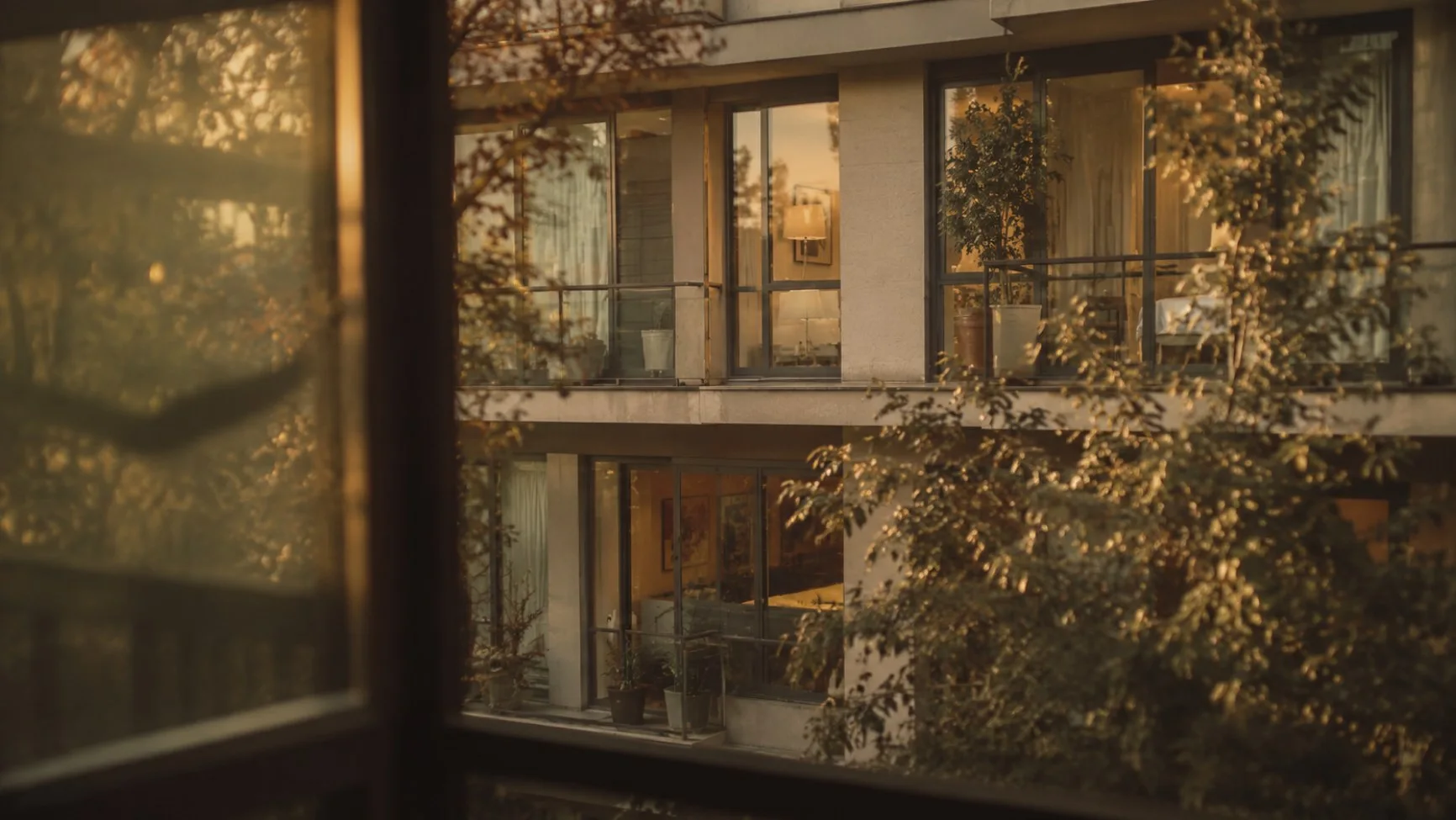 A community's lit balconies and courtyard trees at golden hour, seen through a window.